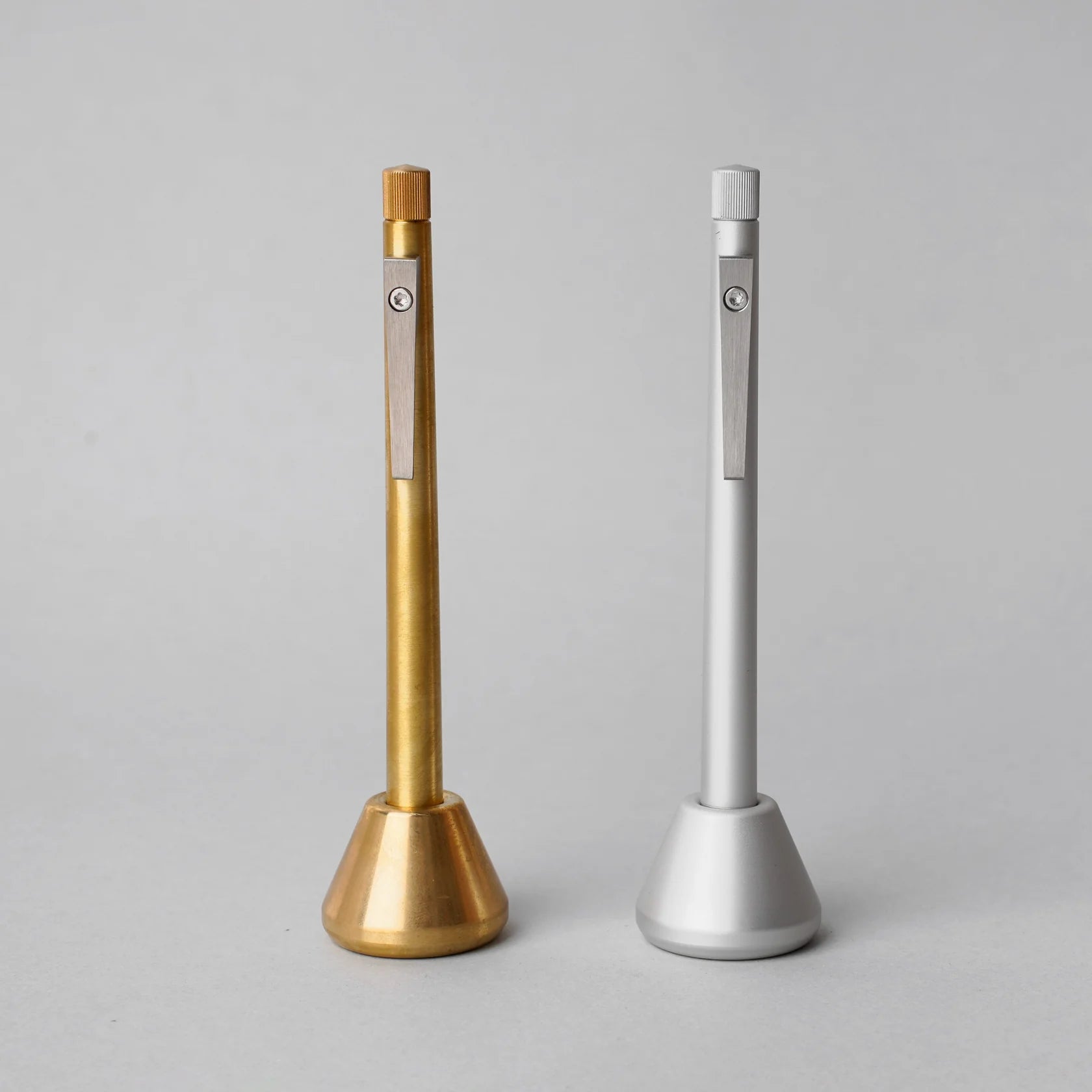 Two minimalist metal pens displayed upright in matching weighted desk stands, one in brushed brass and one in matte silver, featuring clean cylindrical forms, knurled end details, and precision-machined clips against a neutral gray background.