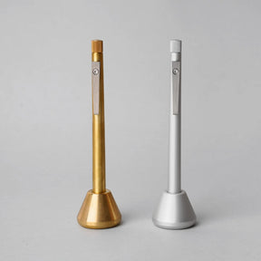 Two minimalist metal pens displayed upright in matching weighted desk stands, one in brushed brass and one in matte silver, featuring clean cylindrical forms, knurled end details, and precision-machined clips against a neutral gray background.