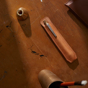Top-down view of a leather pen sheath holding a slim metal pen, placed on a worn wooden desk alongside brass desk accessories, showing natural materials, warm tones, and a handcrafted, minimalist workspace aesthetic.