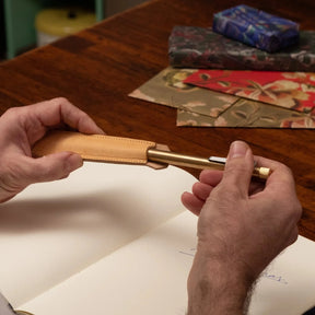Hands holding a brass pen partially inserted into a tan leather pen sheath above an open notebook on a wooden desk, with patterned paper items in the background, highlighting a handcrafted writing accessory in use.