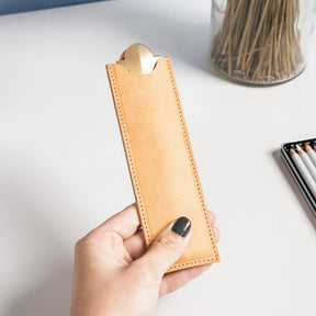 Hand holding a tan leather pencil case on a light surface with a jar of pencils in the background.