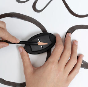 Top-down view of hands using a Blackwing × Makers Cabinet Høvel pencil plane to sharpen a black pencil directly on paper with bold, flowing ink lines.
