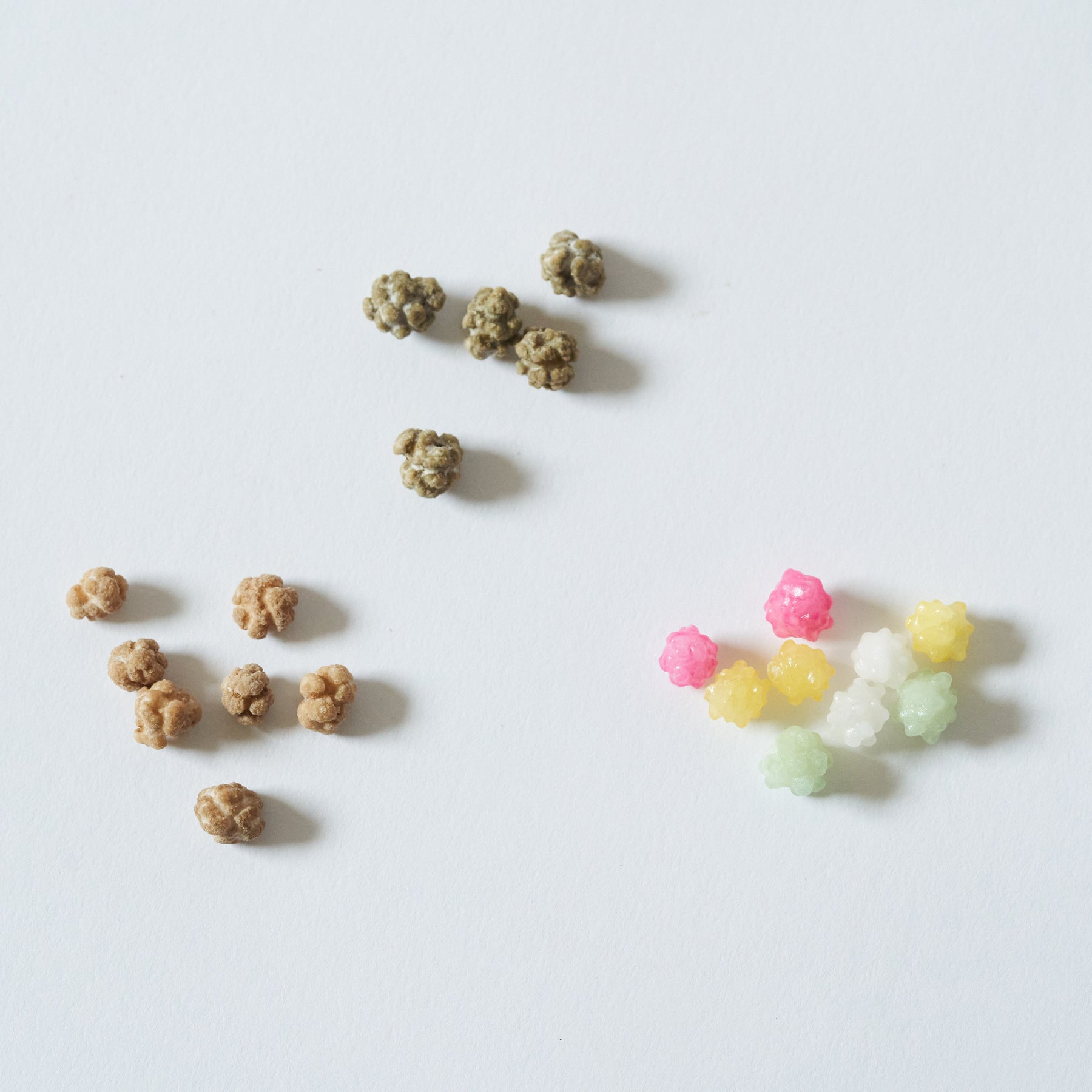 Two small piles of textured pet chews or treats on a plain white background, with one pile in natural tan/brown and the other in pastel pink, yellow, white, and light green.