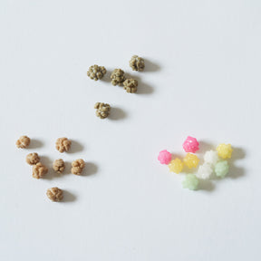 Two small piles of textured pet chews or treats on a plain white background, with one pile in natural tan/brown and the other in pastel pink, yellow, white, and light green.
