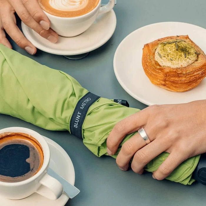 Person holding a green umbrella with 'Blunt Vienna' branding, next to two cups of coffee and a pastry on a table.