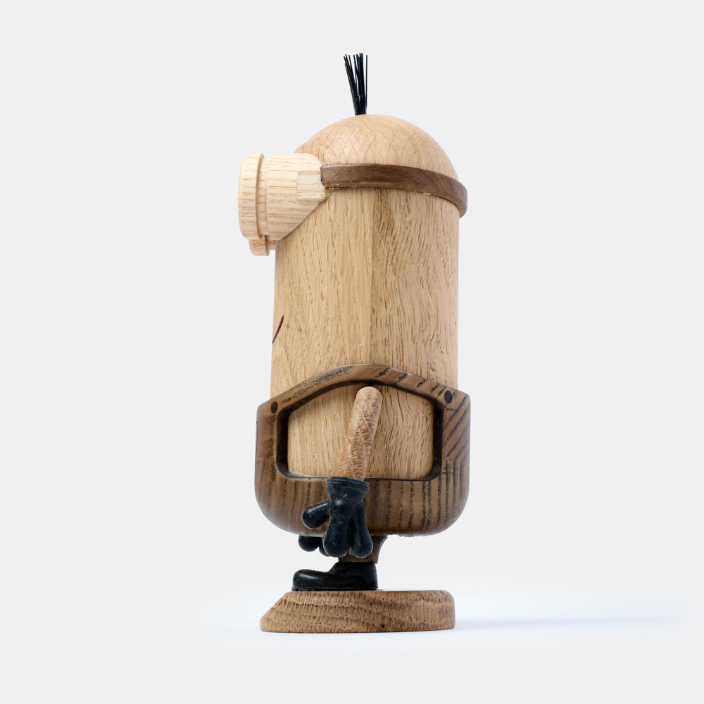 Side view of a wooden figurine of an animated cartoon character carved from light and dark wood, featuring a tall cylindrical body with a single goggle detail, rounded head with small hair detail, sculpted arm with dark glove, visible natural wood grain, and a matte surface on a plain white background.