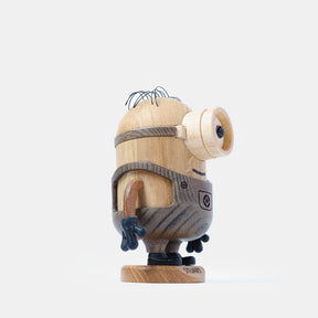 Wooden figurine of an animated cartoon character carved from light and dark wood, featuring a rounded cylindrical body with a single goggle detail, engraved belt and clothing elements, small sculpted arms and legs, visible natural wood grain, and a matte surface on a plain white background.