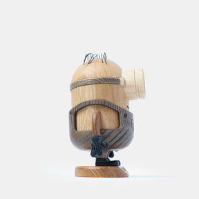 Wooden figurine of an animated cartoon character carved from light and dark wood, featuring a rounded cylindrical body with a single goggle detail, engraved belt and clothing elements, small sculpted arms and legs, visible natural wood grain, and a matte surface on a plain white background.