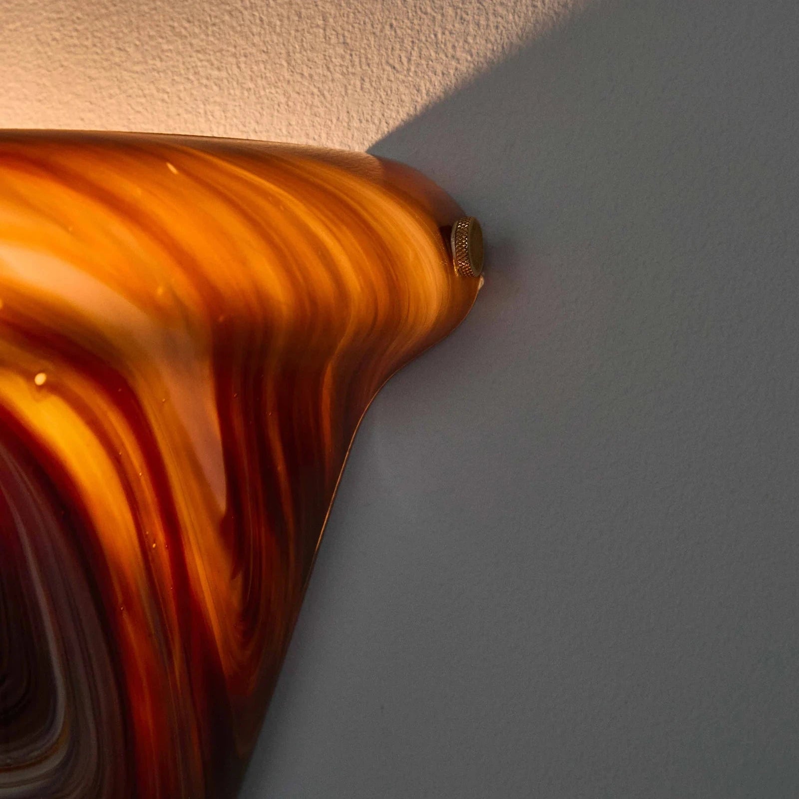 A close-up of a glossy amber and orange marbled glass object against a light gray wall, with warm light reflecting off its curved surface.