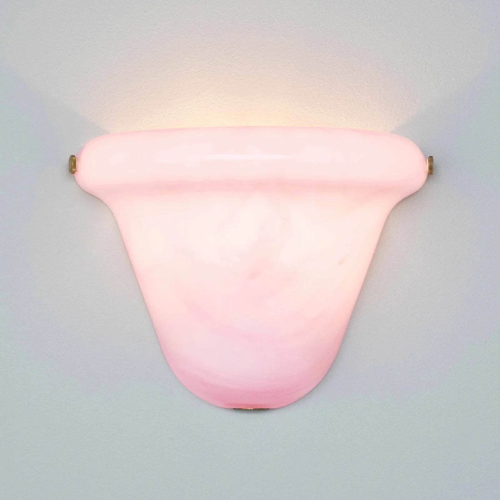 A pink glass wall sconce with a curved, shield-like shade and brass hardware, mounted on a light gray wall and casting a warm glow upwards.