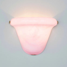 A pink glass wall sconce with a curved, shield-like shade and brass hardware, mounted on a light gray wall and casting a warm glow upwards.