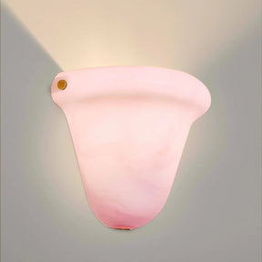 A modern wall sconce light fixture with a curved, bell-shaped translucent pink shade, shown illuminated against a neutral gray wall.