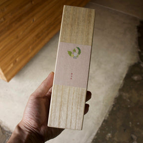A hand holding a tall, narrow rectangular box with a light wood-grain finish and a beige paper band label featuring a small green leaf logo, set against a rustic concrete and wood background.