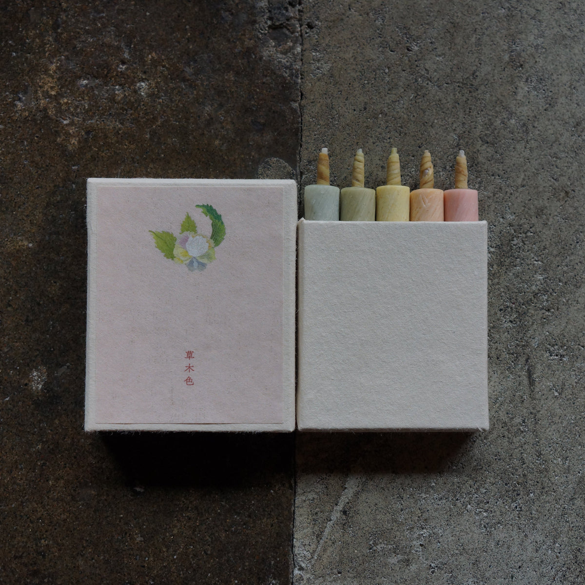 A pair of textured matchboxes on a dark split background. The left matchbox is closed with a pale pink cover featuring a small green and white floral illustration and red characters.