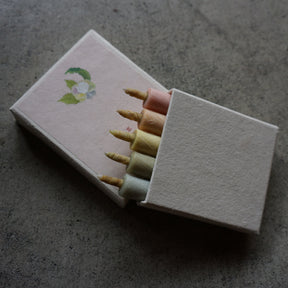 A matchbox-style candle holder with five wax candles inserted, photographed on a textured gray surface. The box is off-white with a small green and blue floral illustration on the lid, and the visible wax colors include pink, yellow, cream, and pale green.