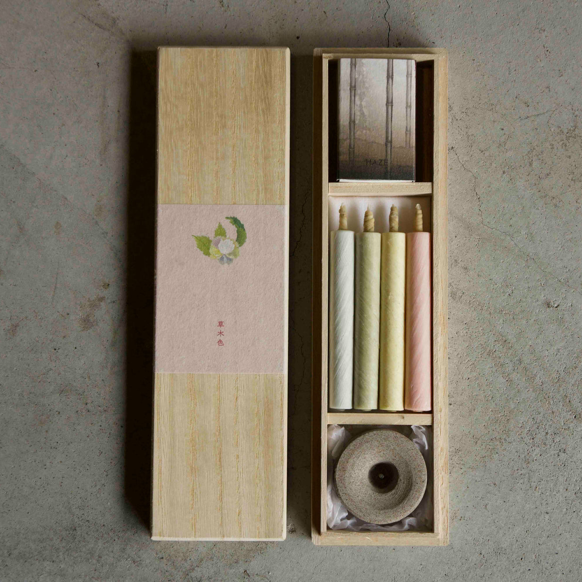 A traditional Japanese incense set in a light wood box, featuring a light wood outer box with a pink paper band and a green leaf illustration, and an inner box containing three incense sticks in cream, yellow, and pink, a roll of incense paper, and a metal incense holder.