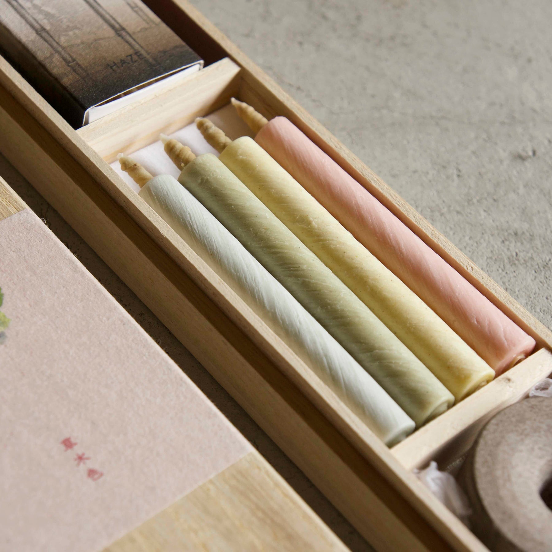 A close-up of a wooden presentation box containing several pastel-colored wax candles in white, pale yellow, and soft pink. The box is shown open, with a matching beige paper label featuring a small green floral illustration and red text on the lid, set against a neutral concrete background.