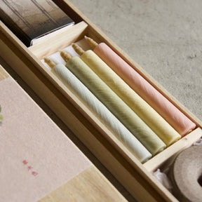 A close-up of a wooden presentation box containing several pastel-colored wax candles in white, pale yellow, and soft pink. The box is shown open, with a matching beige paper label featuring a small green floral illustration and red text on the lid, set against a neutral concrete background.