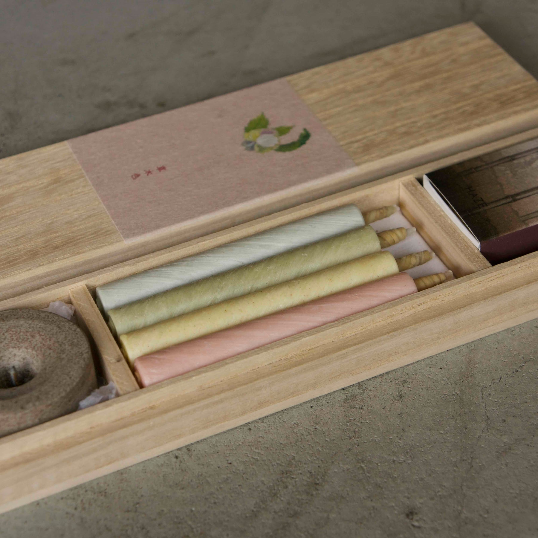 A wooden incense box opened to reveal several rolled incense sticks in pale yellow, cream, and pink tones, a round incense burner, and a small stack of dark rectangular pieces, all arranged on a neutral concrete surface.