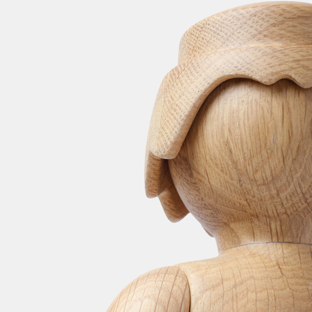 Detail of a wooden Playmobil-style human figure showing a smooth carved leg and curved hand, highlighting the natural light oak wood grain and softly rounded sculptural forms against a plain white background.