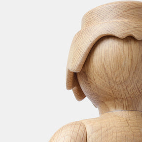 Detail of a wooden Playmobil-style human figure showing a smooth carved leg and curved hand, highlighting the natural light oak wood grain and softly rounded sculptural forms against a plain white background.