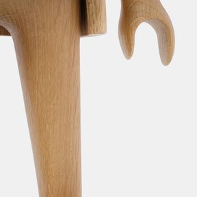 Detail of a wooden Playmobil-style human figure showing a smooth carved leg and curved hand, highlighting the natural light oak wood grain and softly rounded sculptural forms against a plain white background.