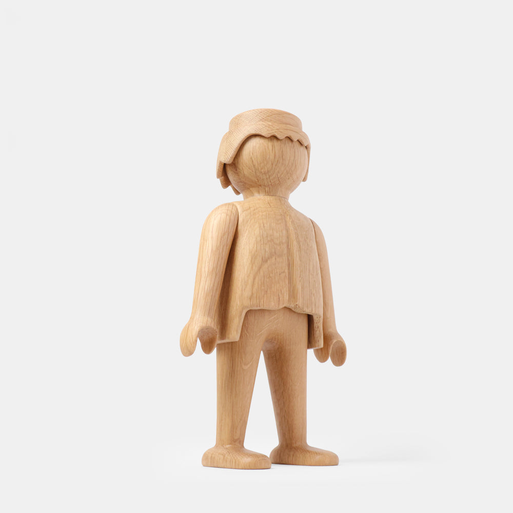 Wooden Playmobil-style human figure made from light oak, featuring simplified proportions, a rounded head with carved hair detail, smooth sculpted arms and legs, visible natural wood grain, and a matte surface on a plain white background.