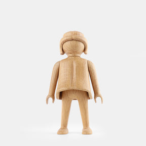 Wooden Playmobil-style girl figure made from light oak, featuring simplified proportions, a rounded head with carved hair detail, smooth sculpted arms and legs, visible natural wood grain, and a matte surface on a plain white background.