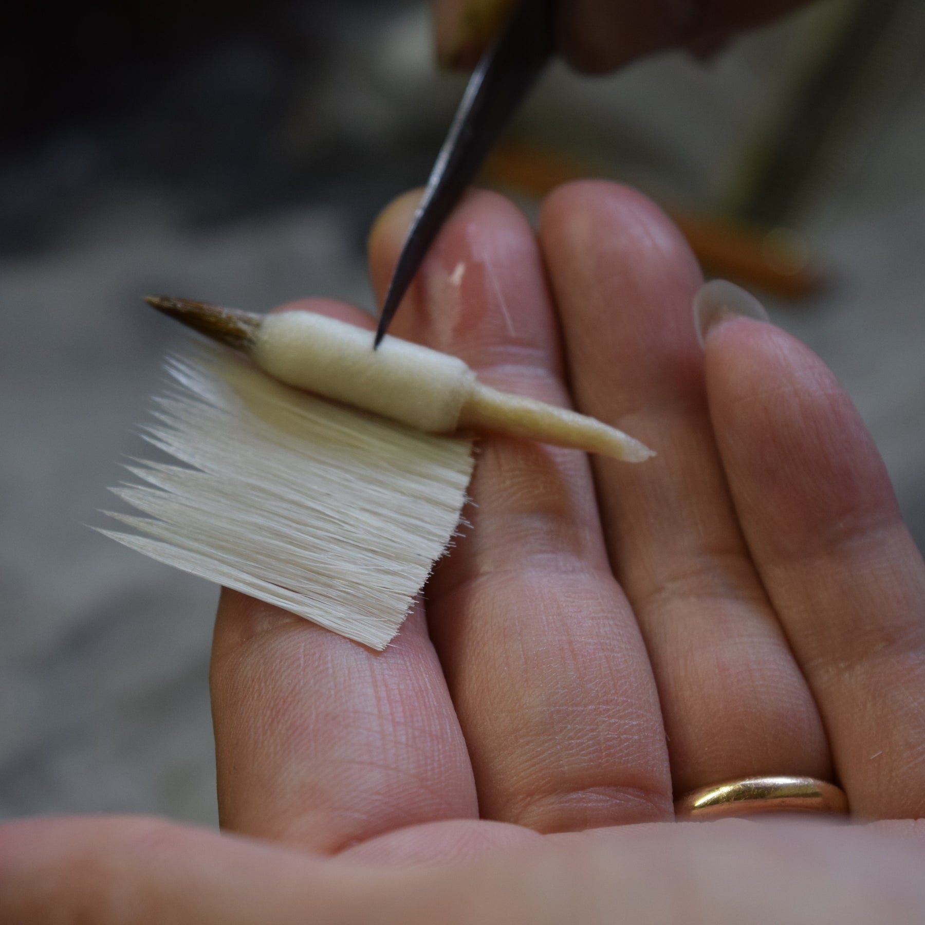 A close-up of a hand holding a small white feather hair accessory with a wooden center, while tweezers adjust the tip. A gold ring is visible on the finger.
