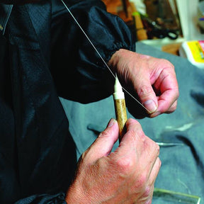 A close-up of a person's hands holding a small gold-colored object and threading a thin string through it, with a dark jacket and a workshop background visible.