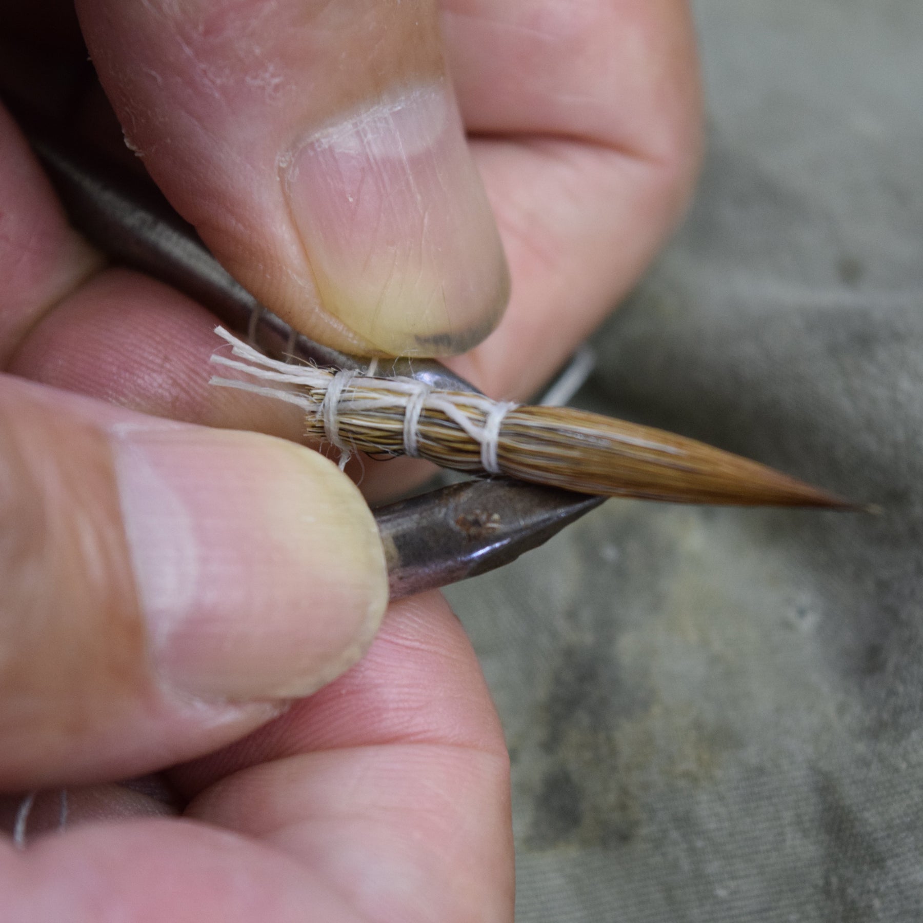 Close-up of a person's hands wrapping a small brown wooden stick or hook with white thread around its middle, against a gray fabric background.