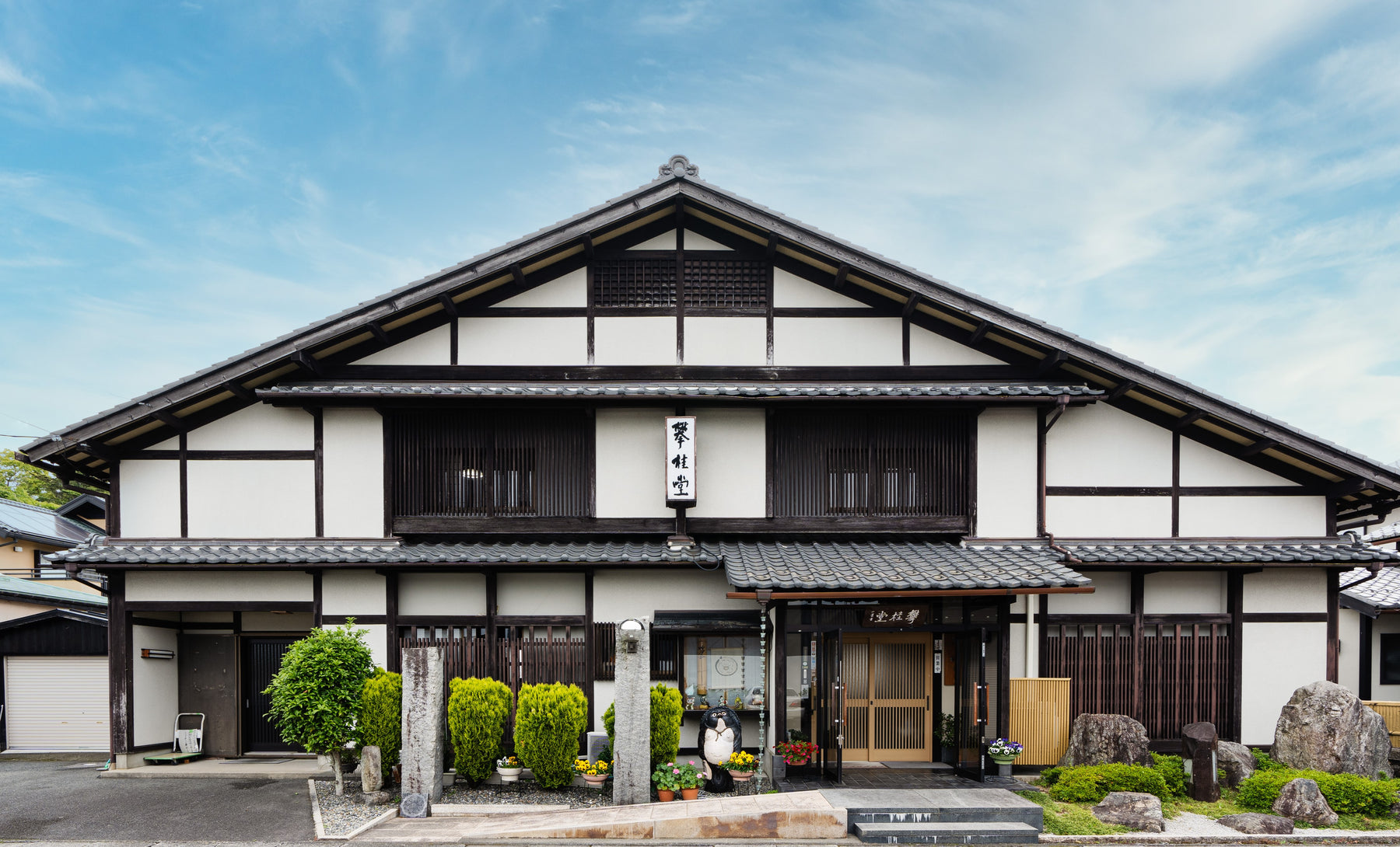 Traditional Japanese wooden building with white walls and dark brown timber framing, featuring a tiled roof, storefront entrance, small garden shrubs, and a blue sky background.