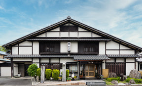 Traditional Japanese wooden building with white walls and dark brown timber framing, featuring a tiled roof, storefront entrance, small garden shrubs, and a blue sky background.