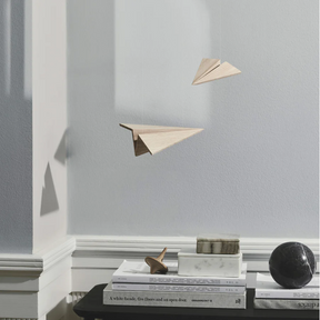 Minimal interior scene featuring two suspended wooden paper-airplane sculptures crafted from light oak, floating against a pale gray wall above a black table stacked with books and small sculptural objects, with soft natural light casting subtle shadows.