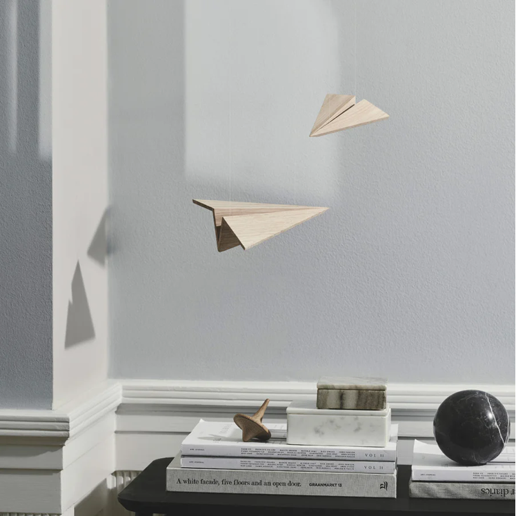 Minimal interior scene featuring two suspended wooden paper-airplane sculptures crafted from light oak, floating against a pale gray wall above a black table stacked with books and small sculptural objects, with soft natural light casting subtle shadows.