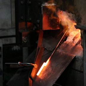 A glowing hot metal ingot or bar held in a furnace or forge, with bright orange and yellow light against dark industrial machinery.