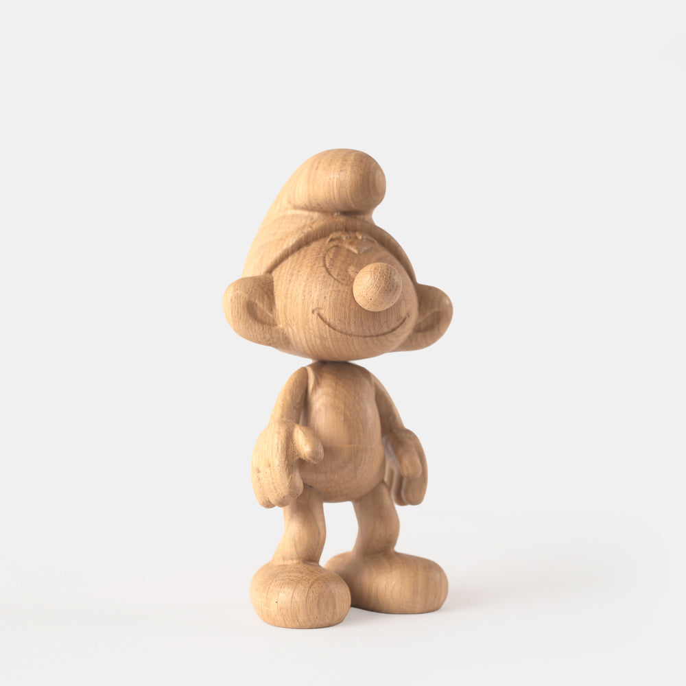 Wooden figurine of a small animated cartoon character, carved from light oak, featuring a rounded hat, smiling facial details, simplified body proportions, visible natural wood grain, and a smooth matte surface on a plain white background.