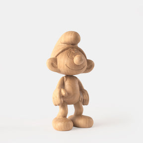 Wooden figurine of a small animated cartoon character, carved from light oak, featuring a rounded hat, smiling facial details, simplified body proportions, visible natural wood grain, and a smooth matte surface on a plain white background.
