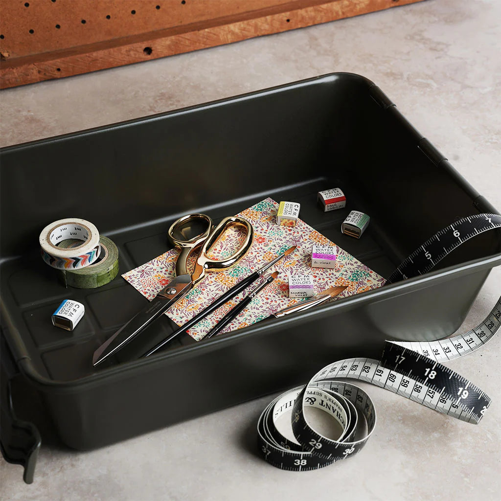 military green storage bin holding craft supplies including scissors, washi tape, paint pans, brushes, patterned paper, and a measuring tape, shown on a work surface.