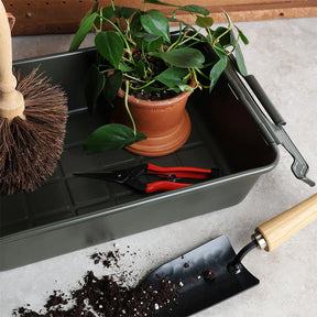 Military green storage bin holding a potted houseplant, pruning shears, and a scrub brush, with potting soil and a hand trowel on a work surface.