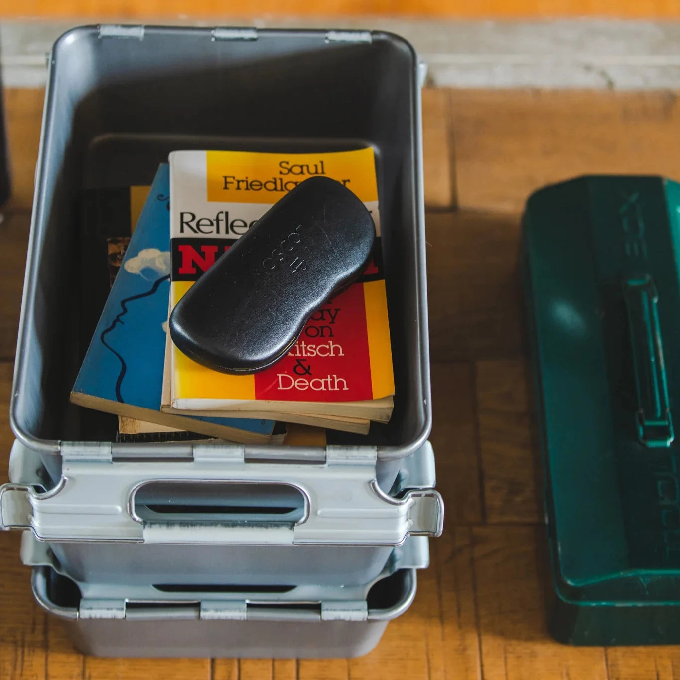 Stackable metal storage bin holding paperback books and a glasses case, placed on a wooden surface.