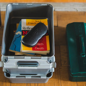 Stackable metal storage bin holding paperback books and a glasses case, placed on a wooden surface.