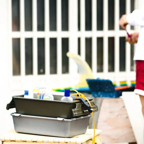 Stacked storage bins holding sunscreen and personal items on a table at a poolside or outdoor setting, with swim toys and a child in the background.