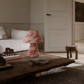 Living room with a floral-patterned lamp on a wooden table, a bed in the background, and a door open to another room.