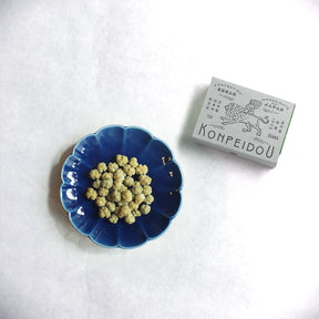 A small blue scalloped dish filled with round pale yellow beads or seeds, placed on a white textured surface next to a gray box labeled KOMPEIDOU.