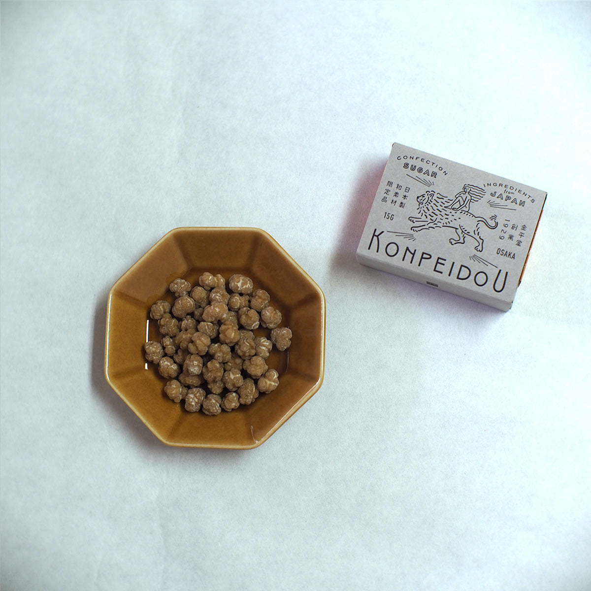 A small hexagonal ceramic dish filled with brown round food pieces sits on a white surface next to a small product box labeled KOMPEIDOU.