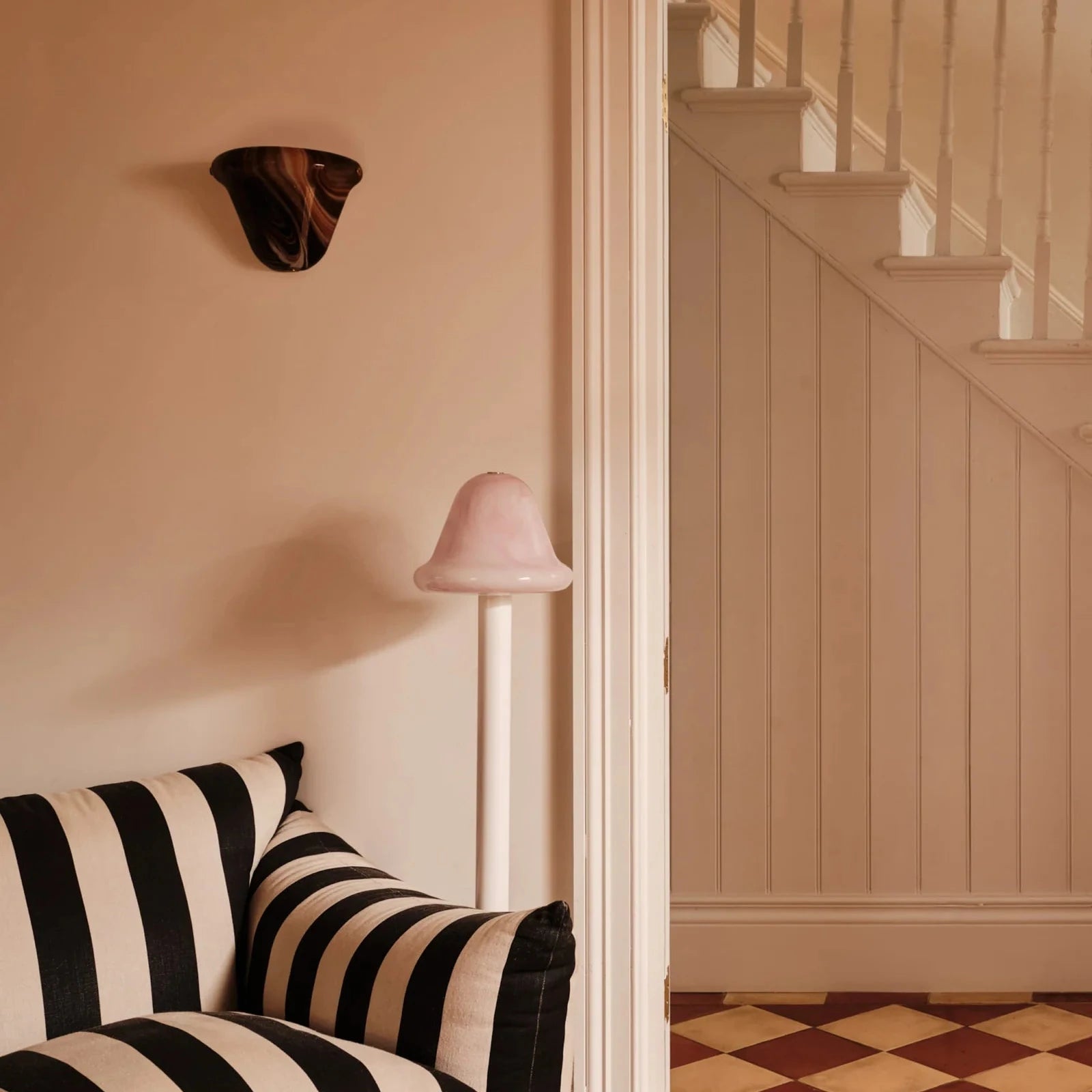 A styled interior corner showing a black and cream striped armchair, a pink floor lamp, a wall sconce, and an open doorway leading to a staircase.