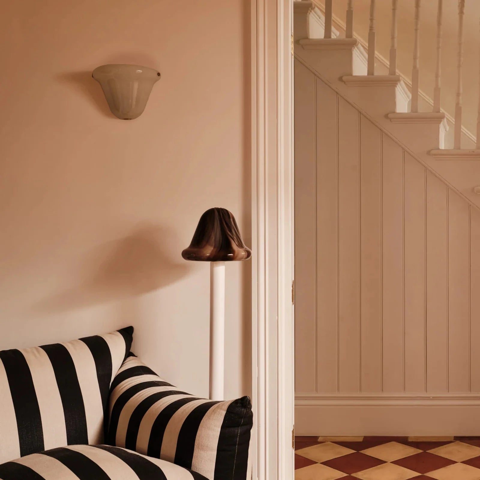 A black and white striped armchair in a warm beige-toned room, with a floor lamp, wall sconce, and an open doorway revealing a staircase.