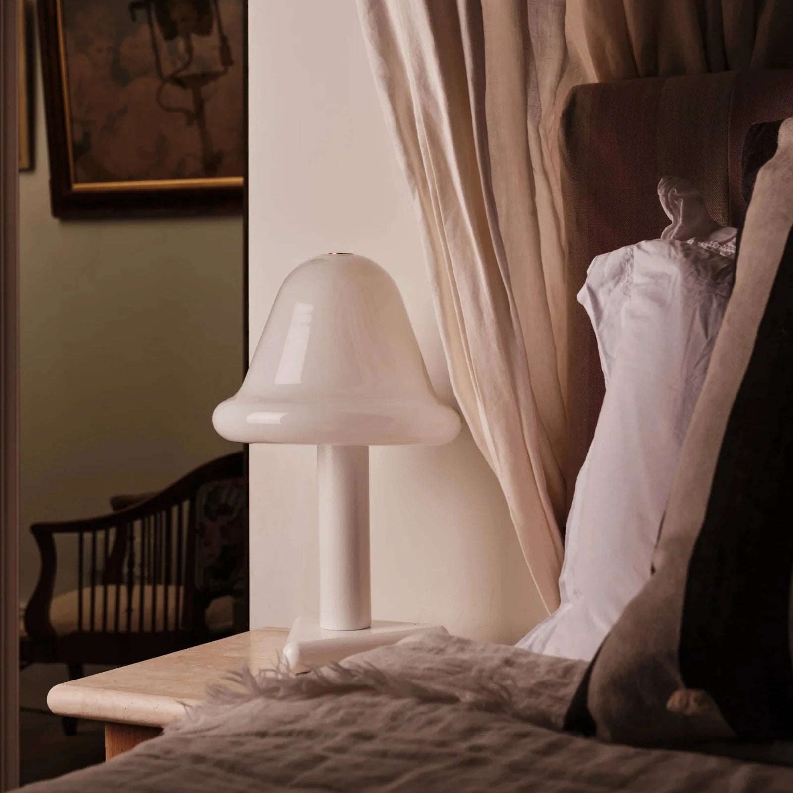 White lamp on a bedside table next to a bed with pillows and curtains.