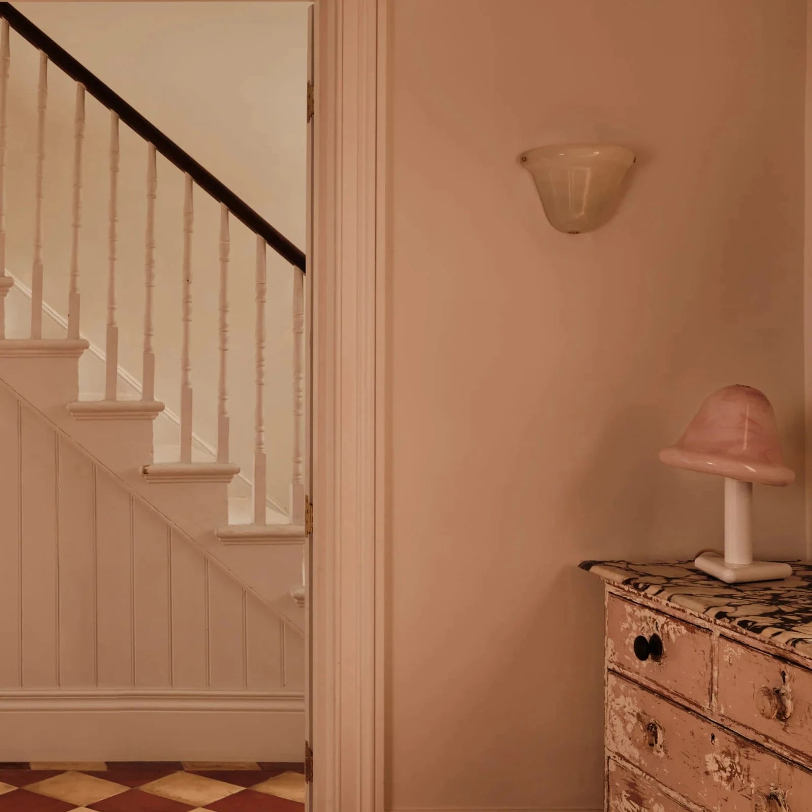 A warm, vintage-style interior scene featuring a dusty pink wall, a white staircase with a dark wooden railing on the left, and a distressed pink and gold chest of drawers with a matching pink lamp on the right.